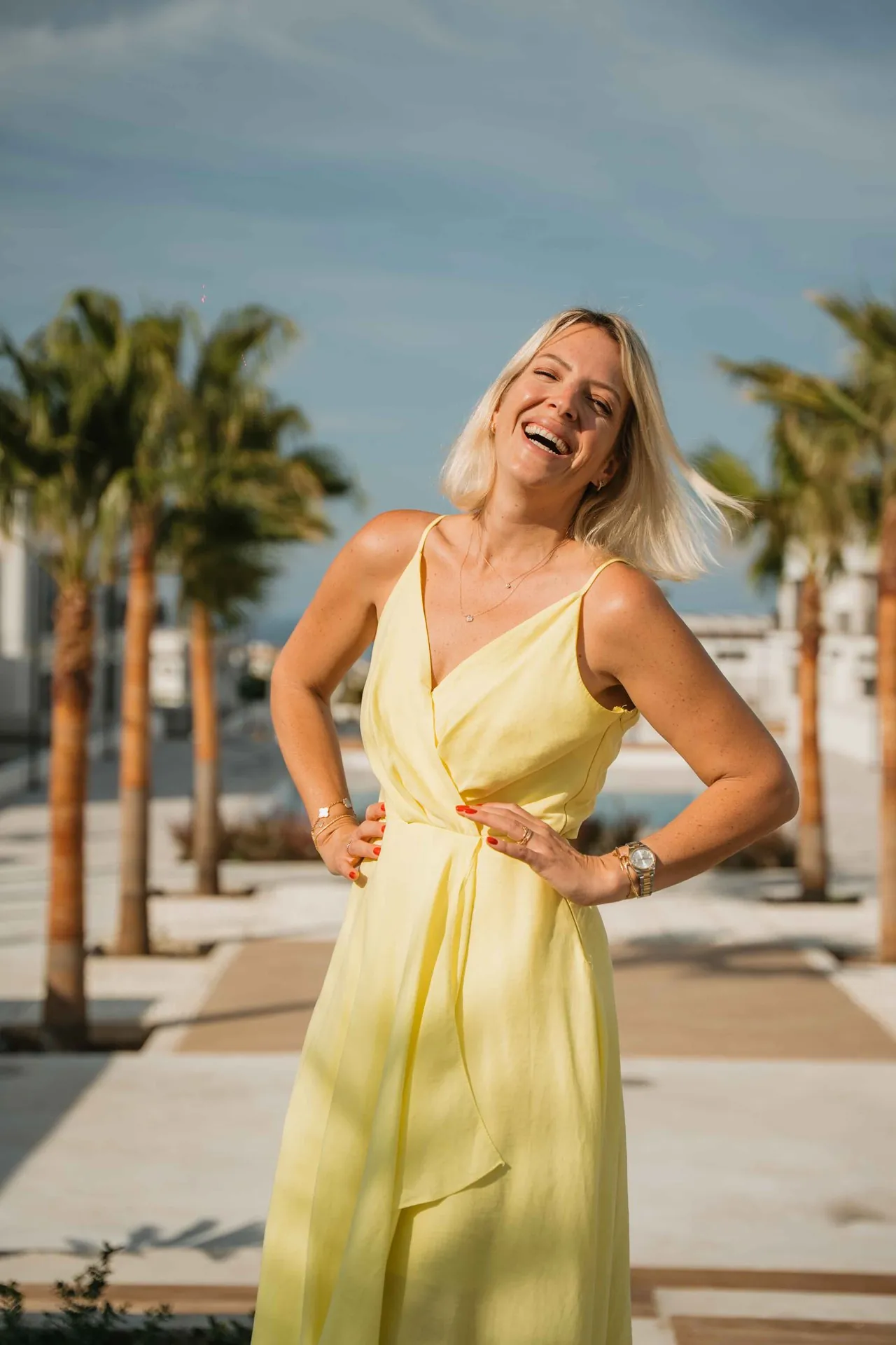 Susanne Rey lacht im gelben Kleid an einer Palmenpromenade